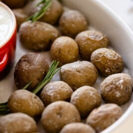 Salt roasted potatoes sitting in white serving platter on bed of salt with sprigs of rosemary and red bowl filled with rosemary Ranch sauce all on white surface