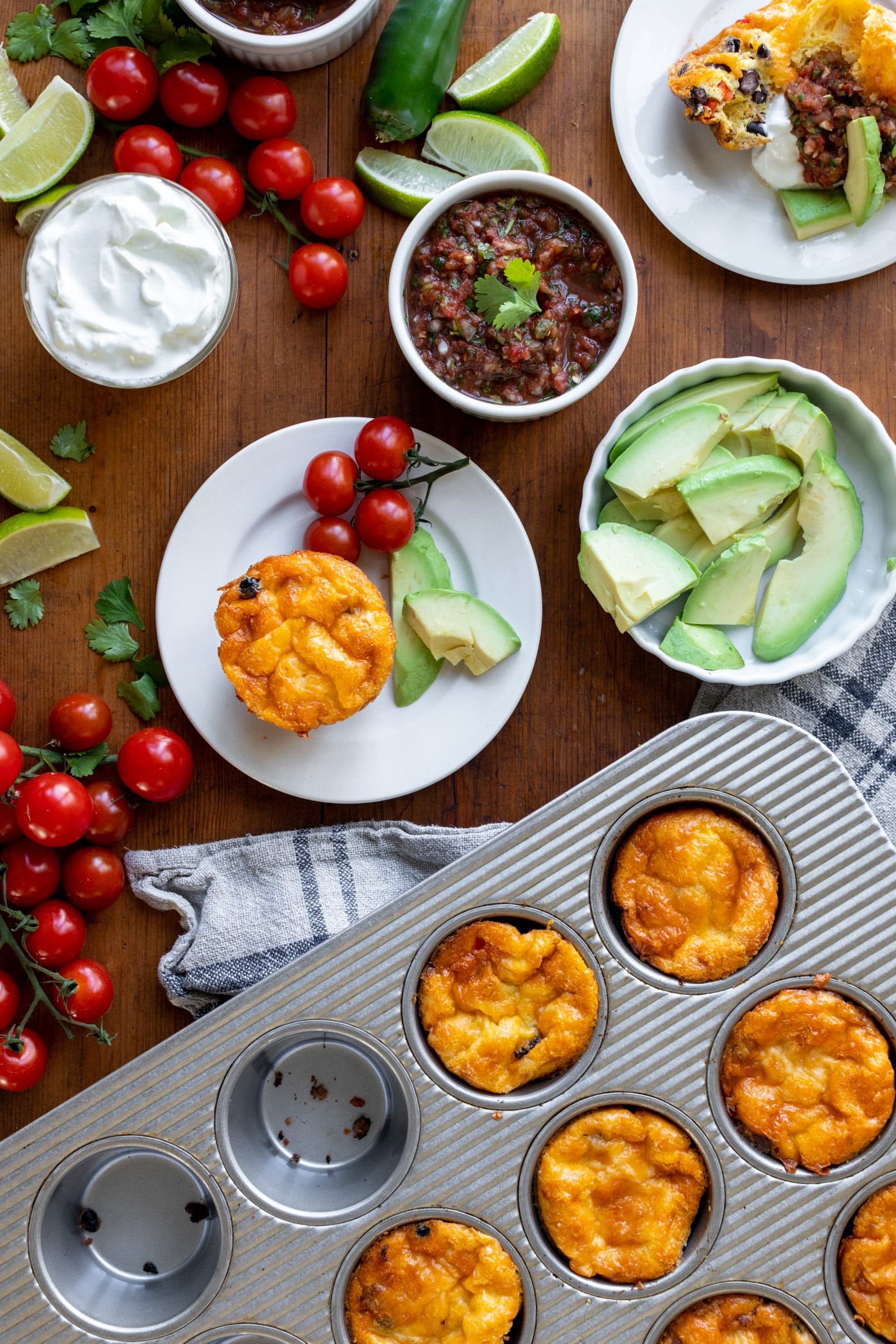 Huevos rancheros egg cups sitting on white plates with pan filled with extra cups as well as many toppings including salsa, sour cream and guacamole all on wood surface