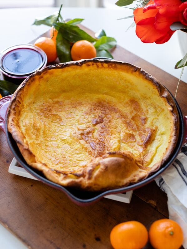 Citrus Dutch baby sitting in a red skillet surrounded by oranges and coulis sauce all on wood board on white surface