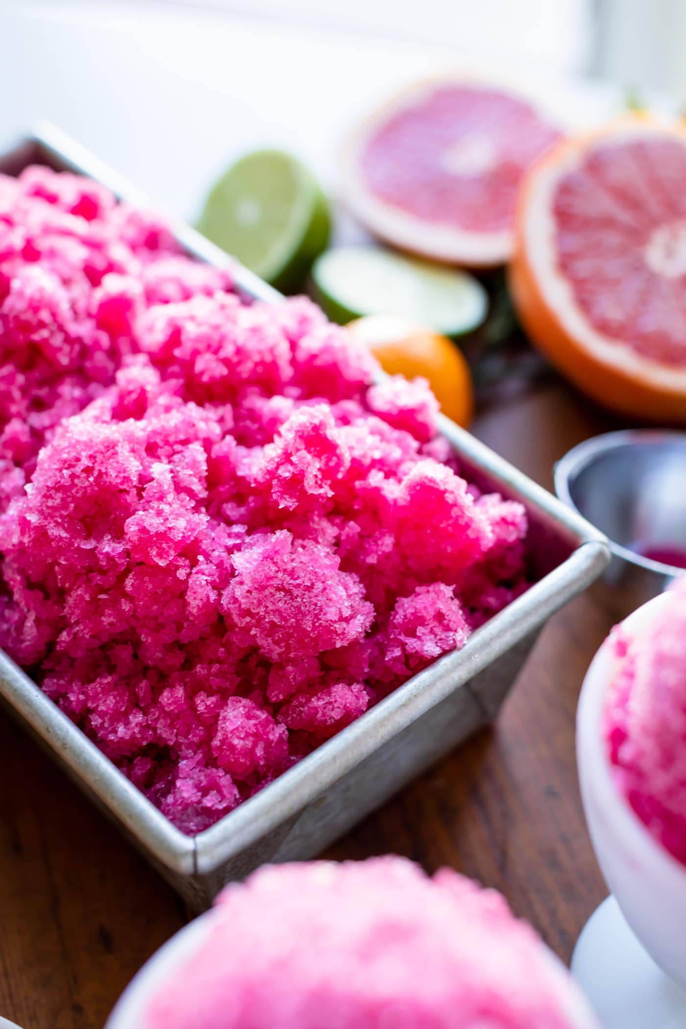 Stainless steel loaf pan filled with grapefruit-flavored pink fruit ice with extra fruit in background all on wood surface