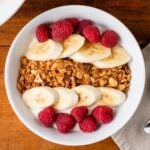 Top down video of homemade peanut butter granola sitting on top of yogurt in a white bowl along with bananas and raspberries with spoons and napkin all on wood surface