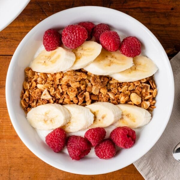 Top down video of homemade peanut butter granola sitting on top of yogurt in a white bowl along with bananas and raspberries with spoons and napkin all on wood surface