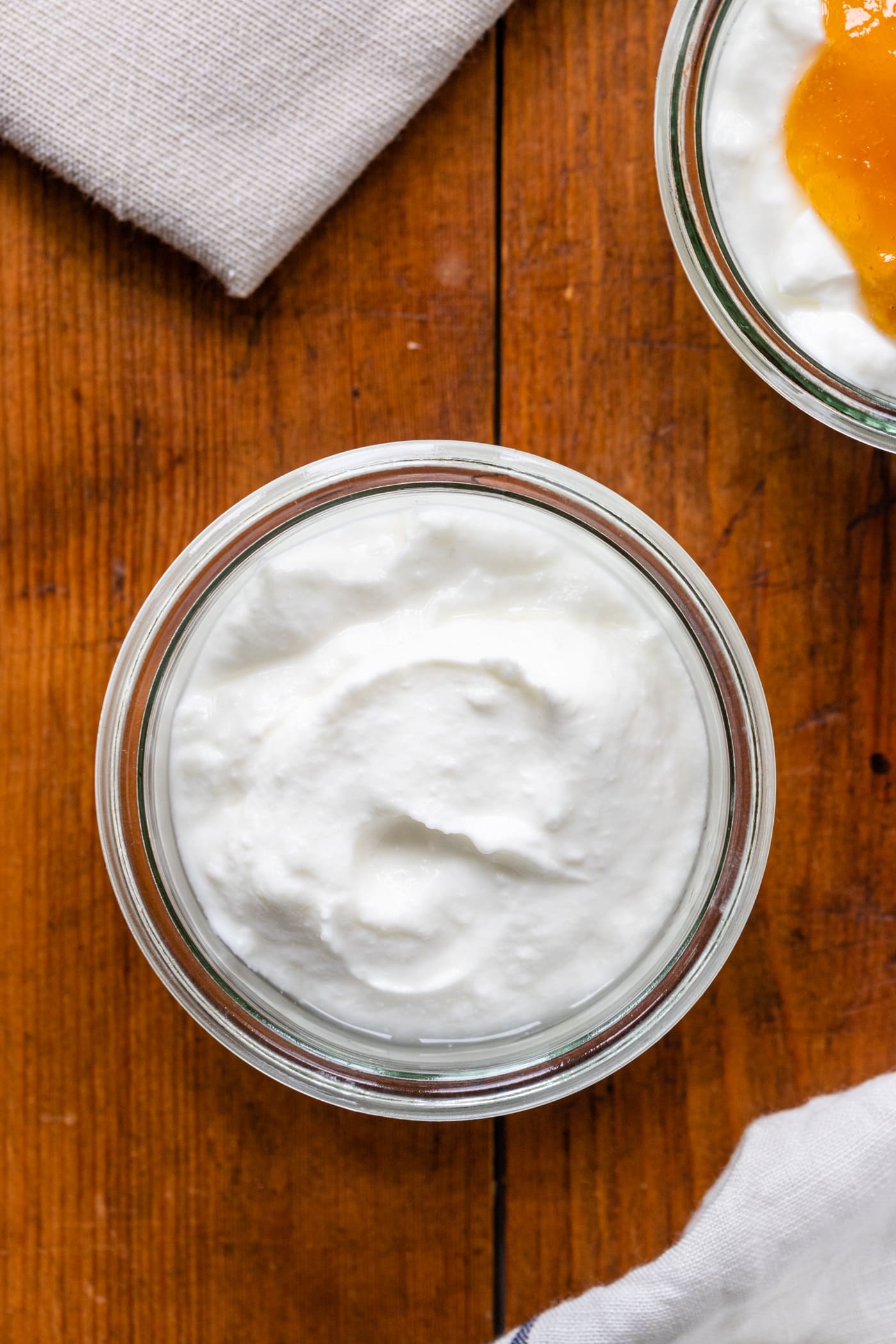 Top down view of yogurt in glass container with no toppings sitting on wood board with towel in corner