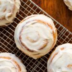 Top down view of ultimate cinnamon roll recipe sitting on wire cooling rack and frosted with cream cheese frosting all on wood surface surrounded by other cinnamon rolls