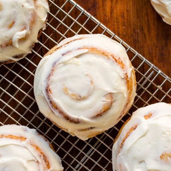 Top down view of ultimate cinnamon roll recipe sitting on wire cooling rack and frosted with cream cheese frosting all on wood surface surrounded by other cinnamon rolls