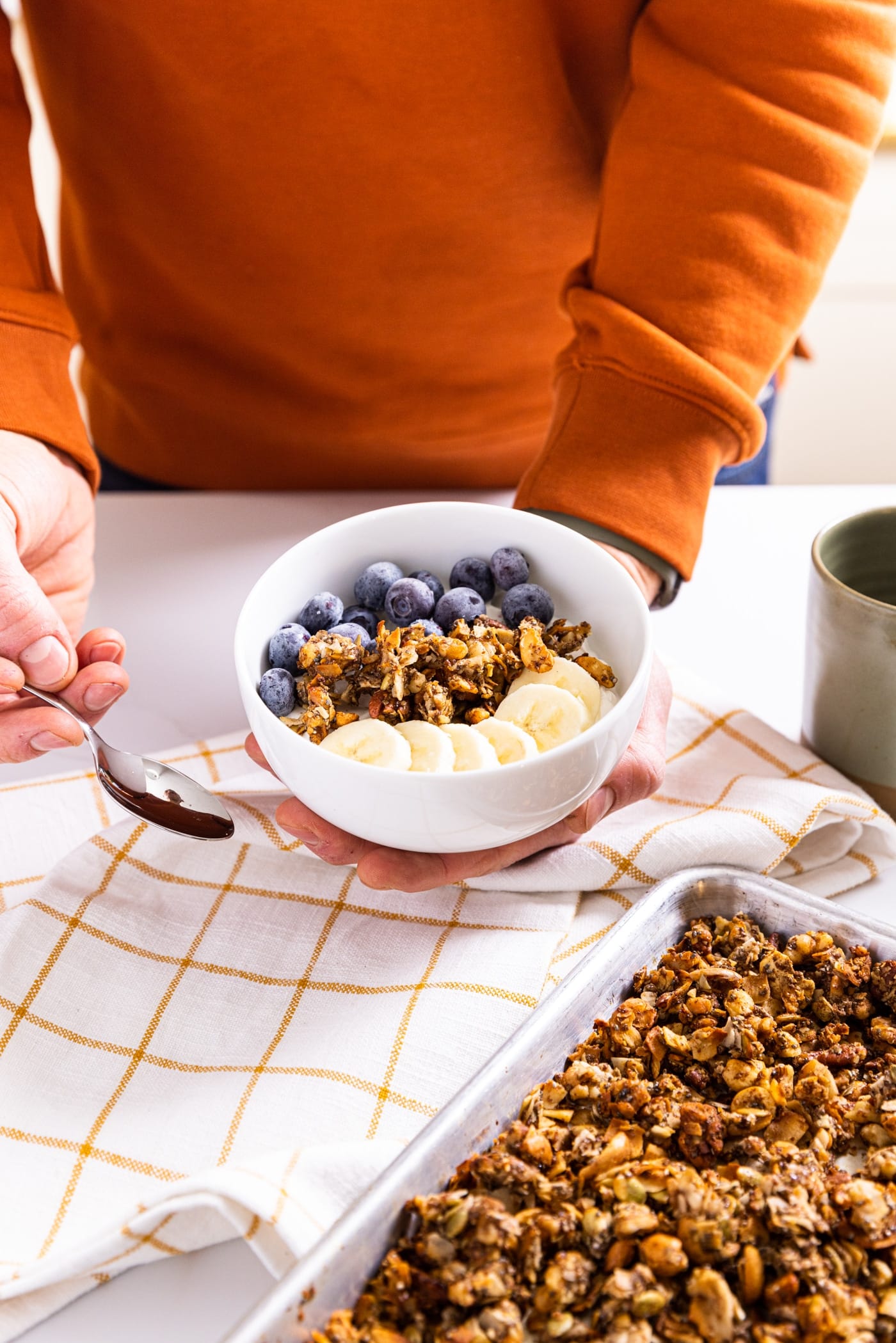 Hand holding bowl and other hand holding spoon filled with grain-free granola on top of yogurt along with blueberries and banana with rest of pan of granola below