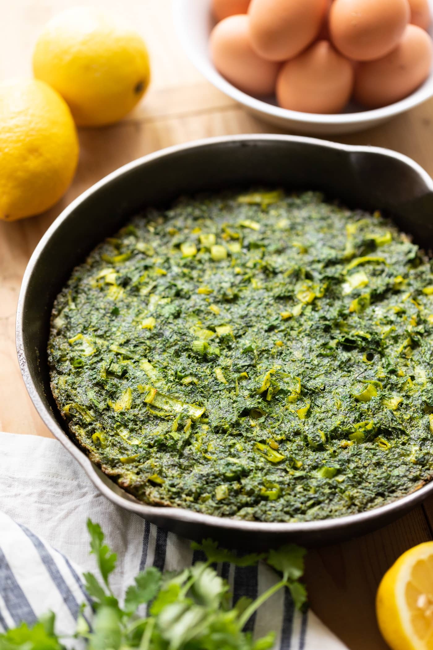 Cast iron skillet filled with omelet dotted with leeks with lemons and eggs in background all on wood surface