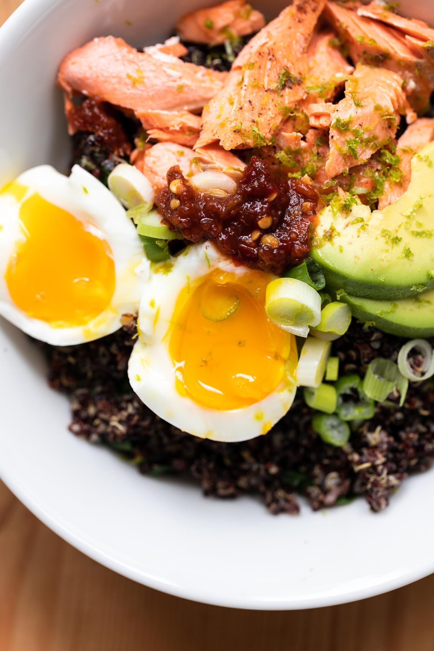 Salmon bowl filled with quinoa, avocado, and sliced open egg all on wood surface