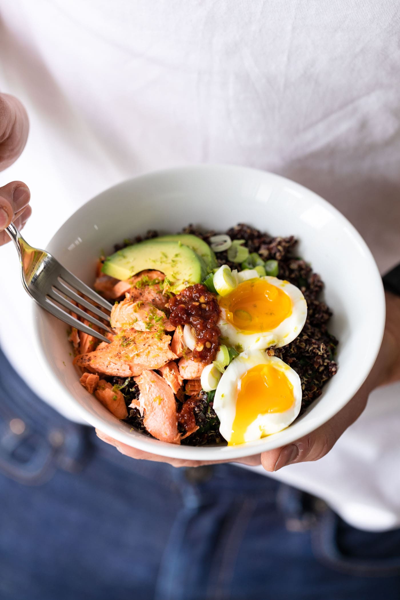 Hands holding white bowl filled with quinoa, avocado, salmon, and a slice egg with a fork taking a piece with white shirt in background