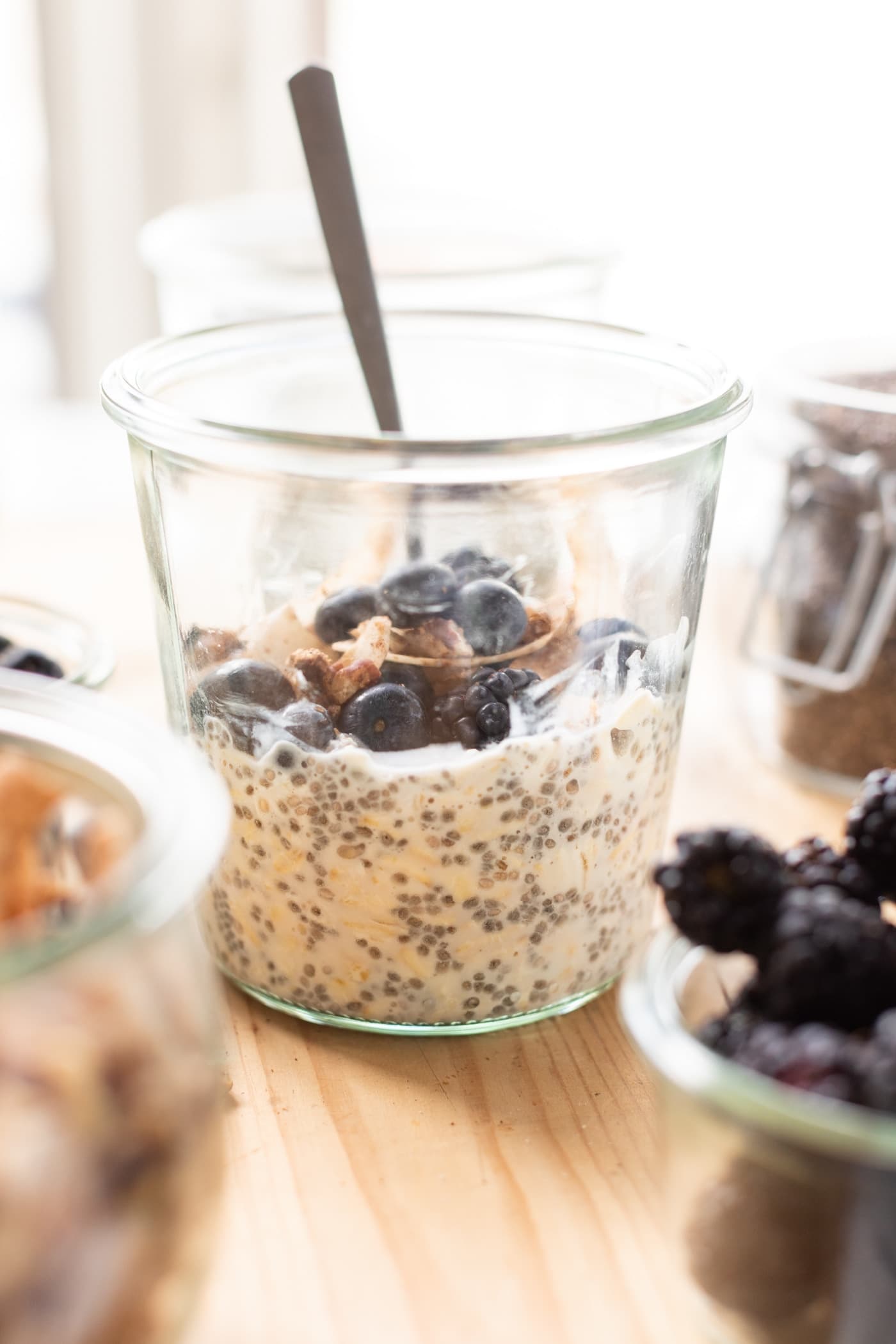 Glass container filled with overnight oats and topped with blueberries and granola with a spoon all on wood surface