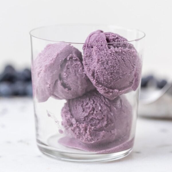 Three scoops of purple blueberry lavender ice cream in glass container with blueberries and ice cream scoop in background all on marble surface