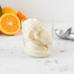 Three scoops of brown butter orange sage ice cream in a glass cup with orange slices and ice cream scoop sitting on piece of marble