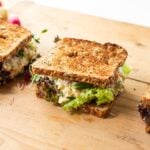 Wood cutting board with three sandwiches made from toasted bread with chickpea salad filling and lettuce with radishes in background