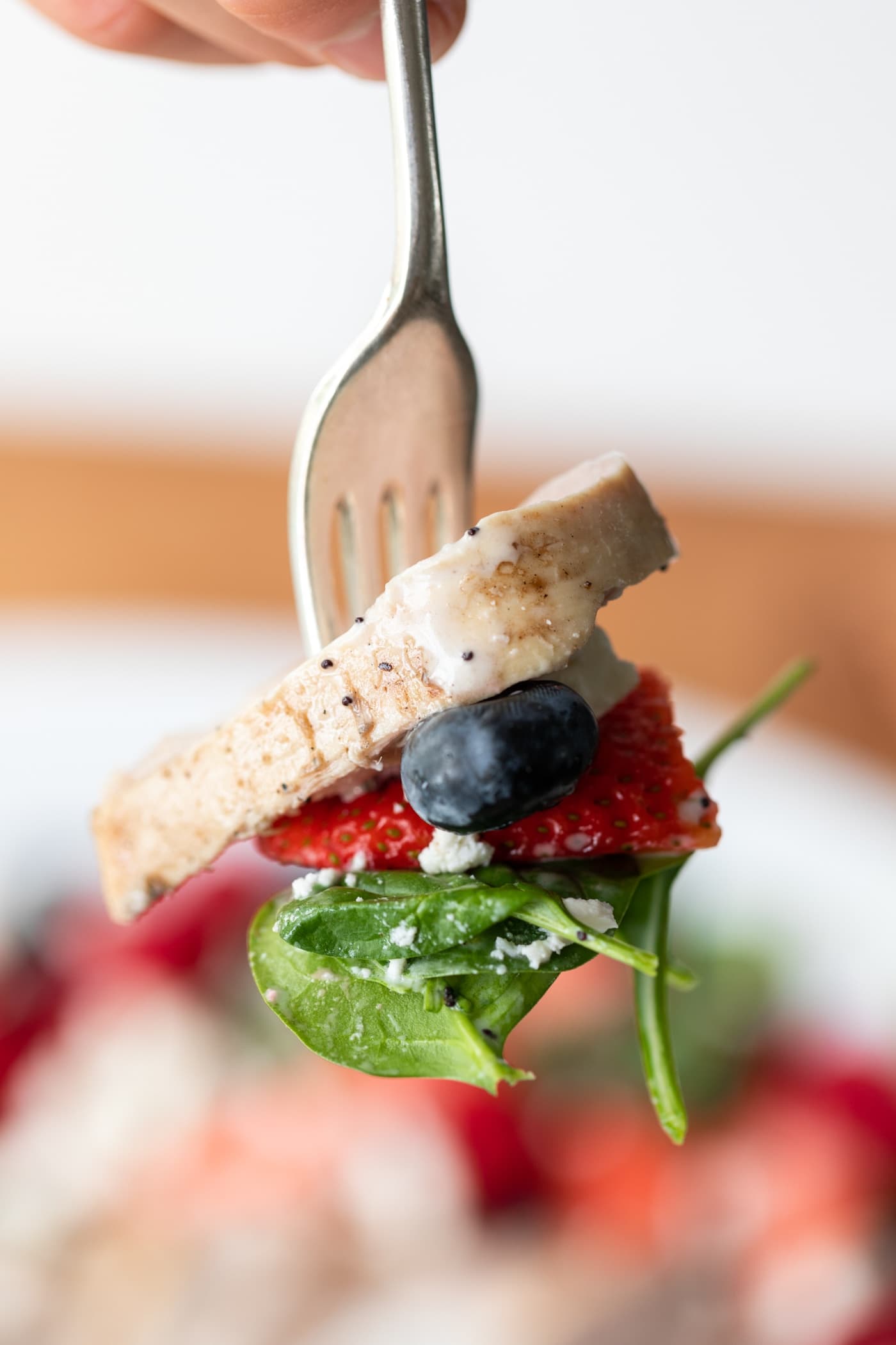 Fork holding piece of chicken as well as blueberry, strawberry, and piece of spinach with salad in background.