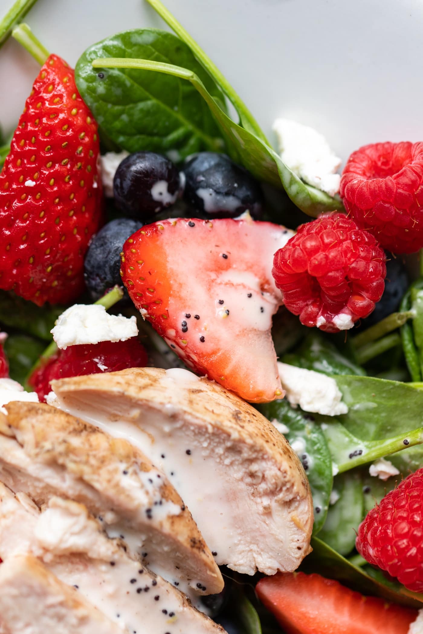 Half piece of strawberry and raspberries and blueberries sitting on spinach with grilled chicken all on white serving platter.