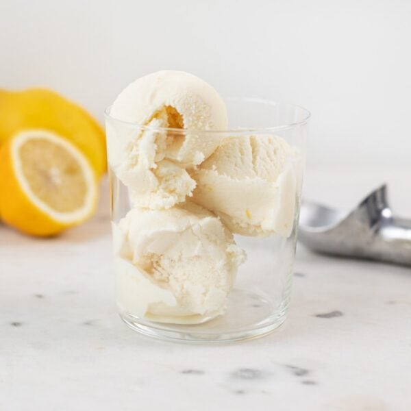 Three scoops of lemon thyme ice cream sitting in glass container with sliced lemon in background and ice cream scoop all on marble surface