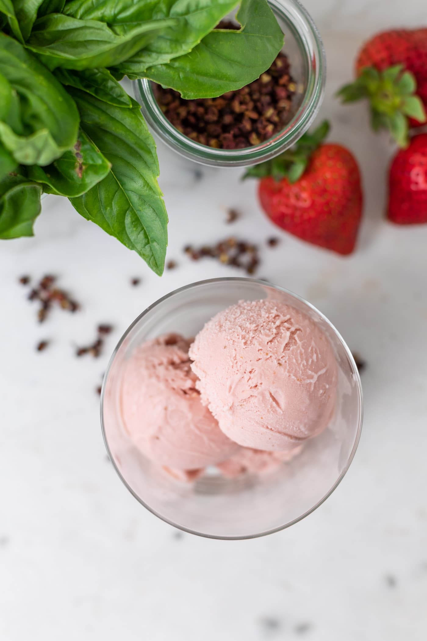 Top down view of pink-colored strawberry basil ice cream in a clear glass cup with basil, peppercorns, and strawberries sitting on marble