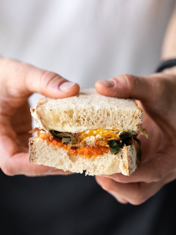 Two hands holding half of harissa egg sandwich topped with Swiss chard with white shirt and jeans in background
