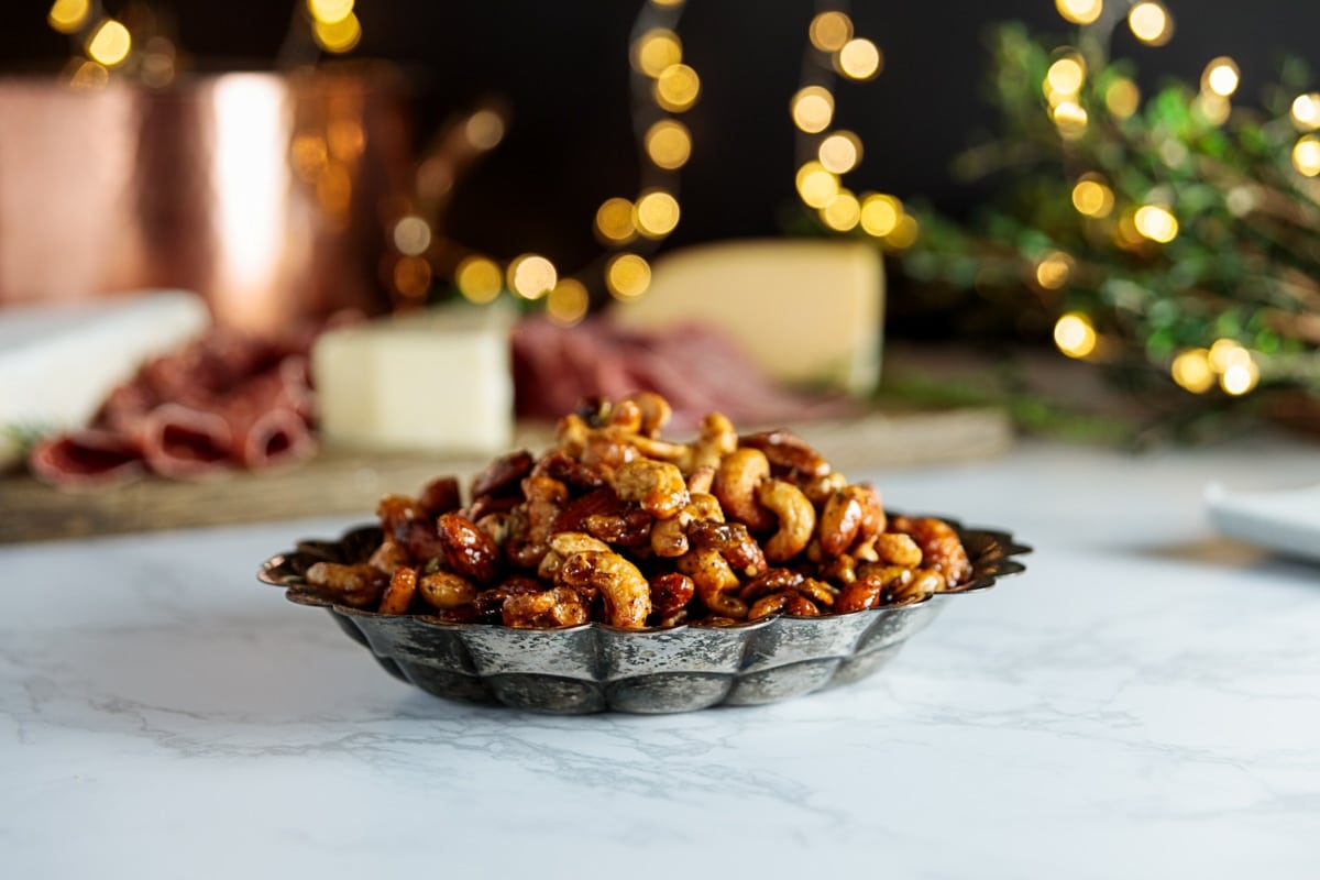Bowl of candied nuts sitting on white piece of marble with twinkling holiday lights in background