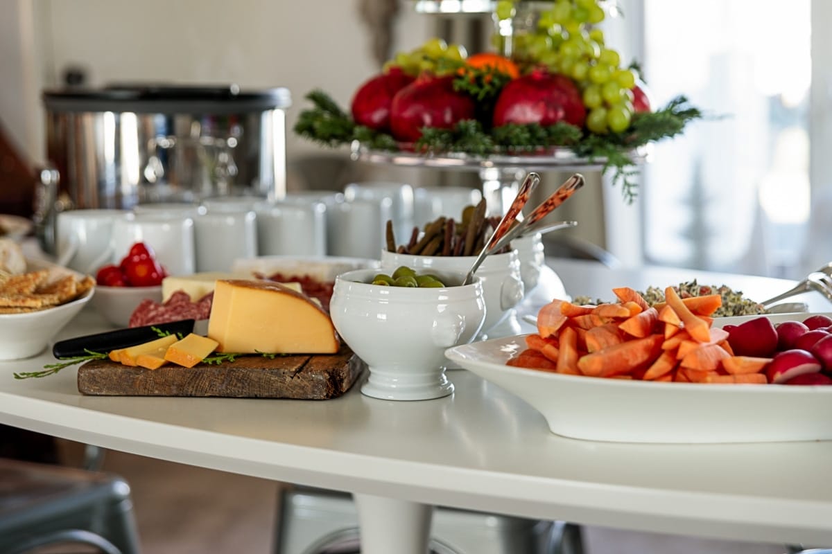 White table filled with dishes for a holiday party including cheese, olives, carrots, fruit, and more, all in white dishes.