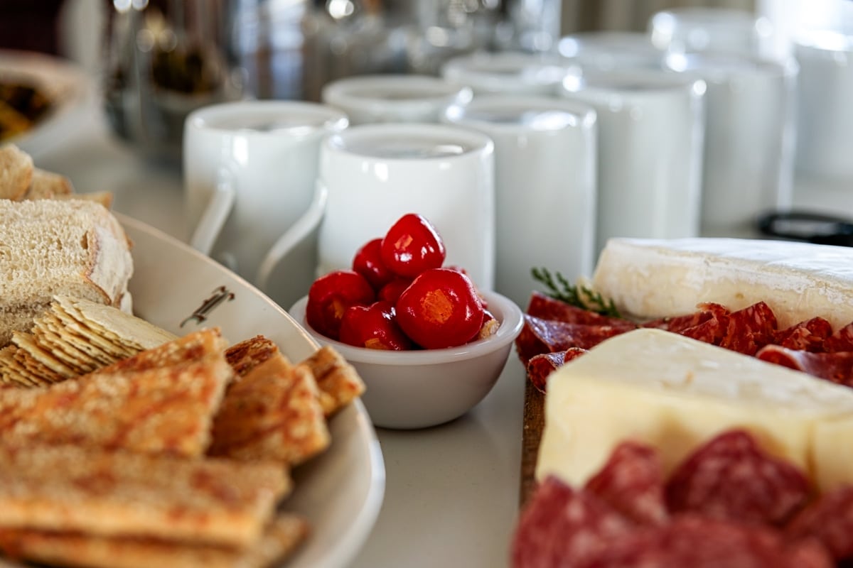 Pickled peppers sitting in small white bowl with white mugs in the background with crackers and cheese and meat in front.