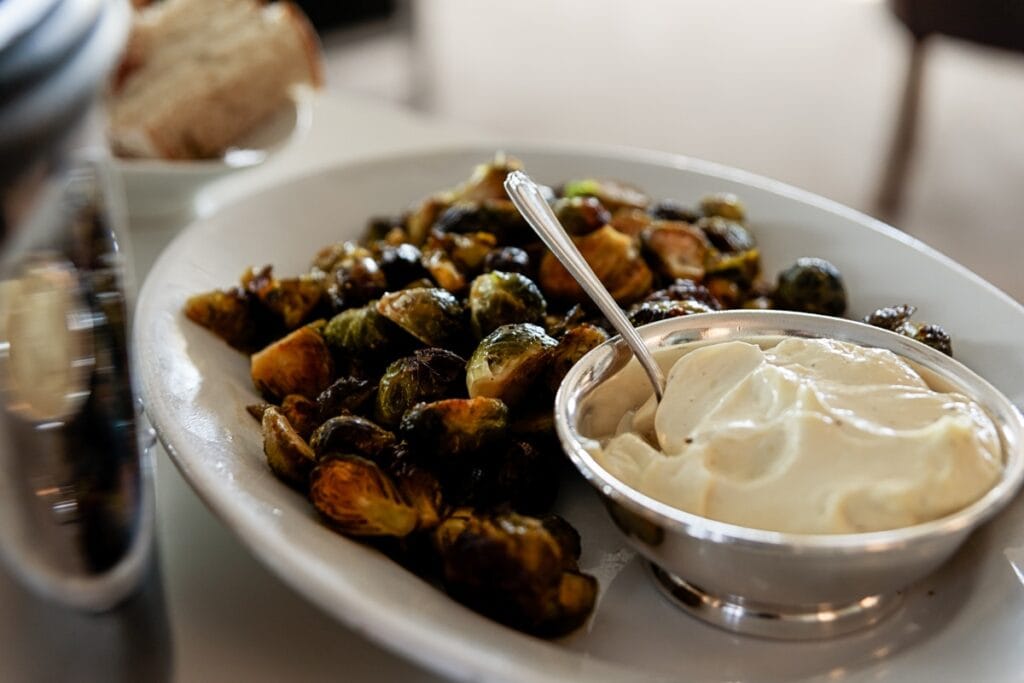 White oval platter with Brussels sprouts and silver container filled with aioli dipping sauce with a spoon.