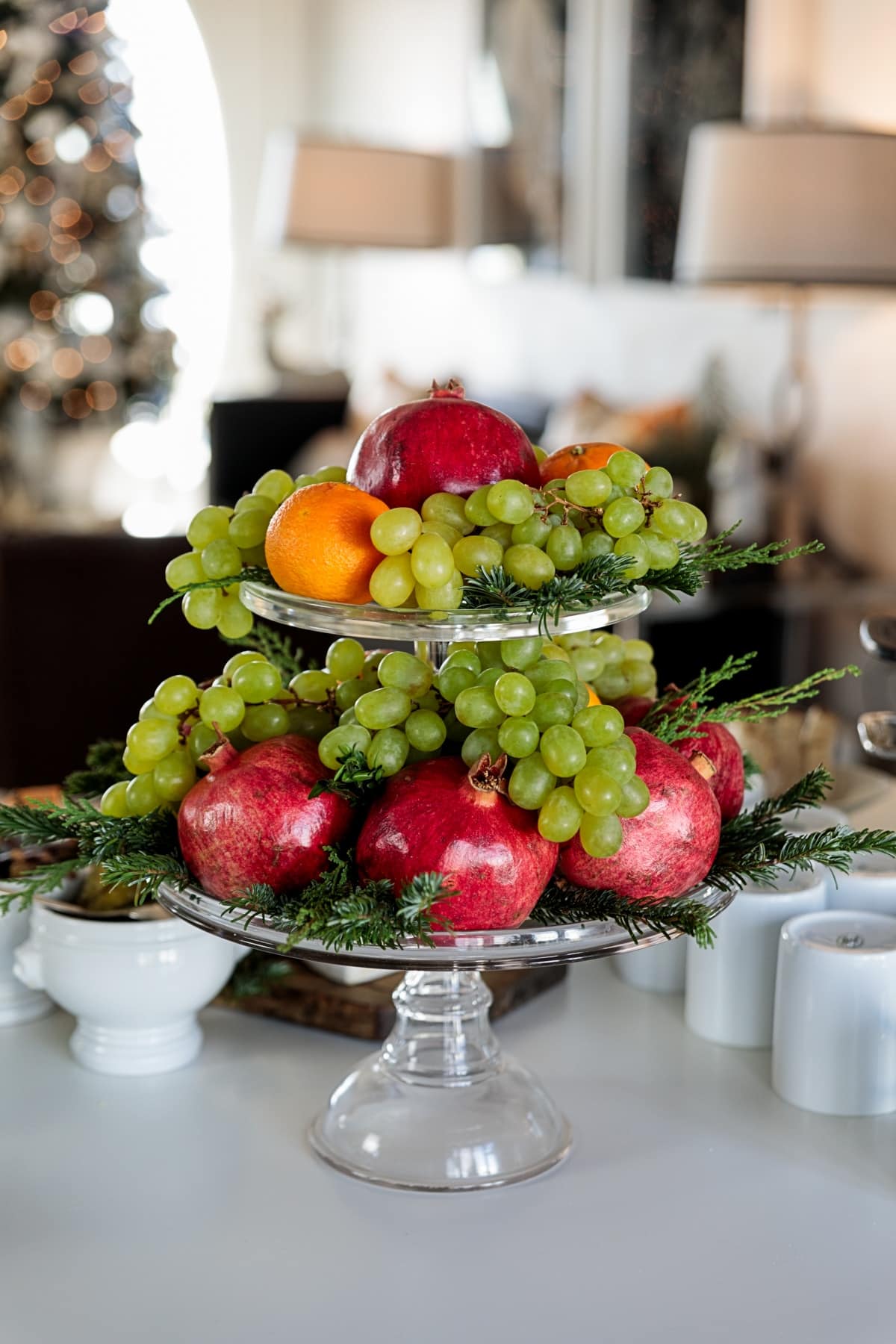 Glass cake stand with multiple levels filled with evergreen with pomegranates, grapes, and oranges all decorating the levels.