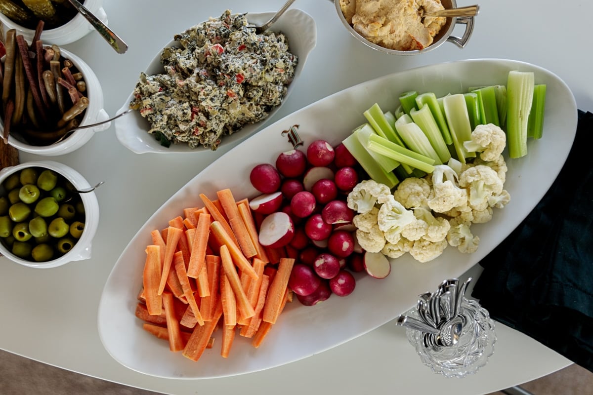 White table filled with dishes for holiday party sides including carrots, celery, radishes, olives, cheese dip, spinach dip, and more.