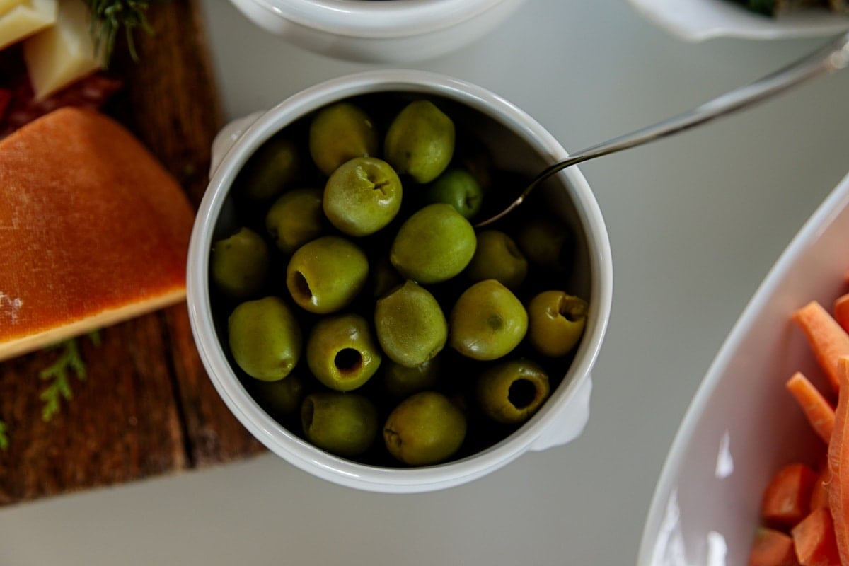 White bowl filled with olives with a spoon on a white table with block of cheese to the side all for a holiday party.