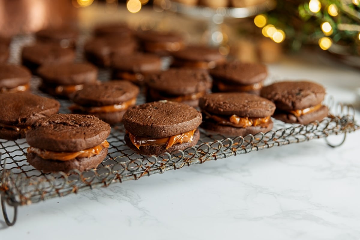 Chocolate sandwich cookies with dulce de leche sauce inside all on cooling rack with twinkling Christmas lights in background.
