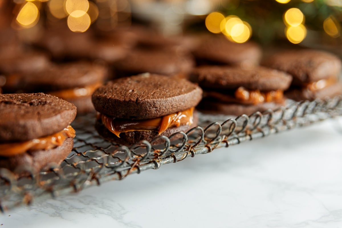 Chocolate sandwich cookie with creamy dulce de leche inside resting with more cookies all on wire cooling rack on white marble surface.