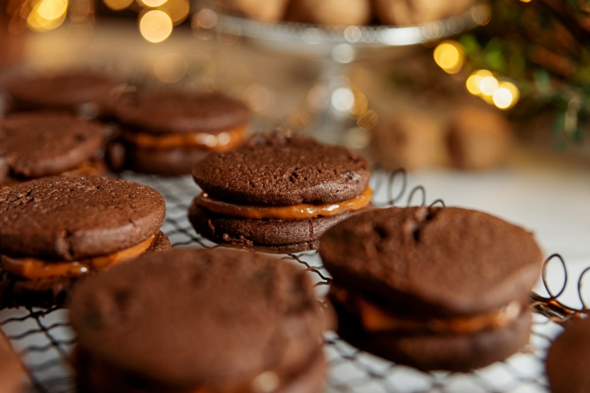 Dulce de leche in between two chocolate cookies creating a sandwich all on wire cooling rack with twinkling Christmas lights in background.