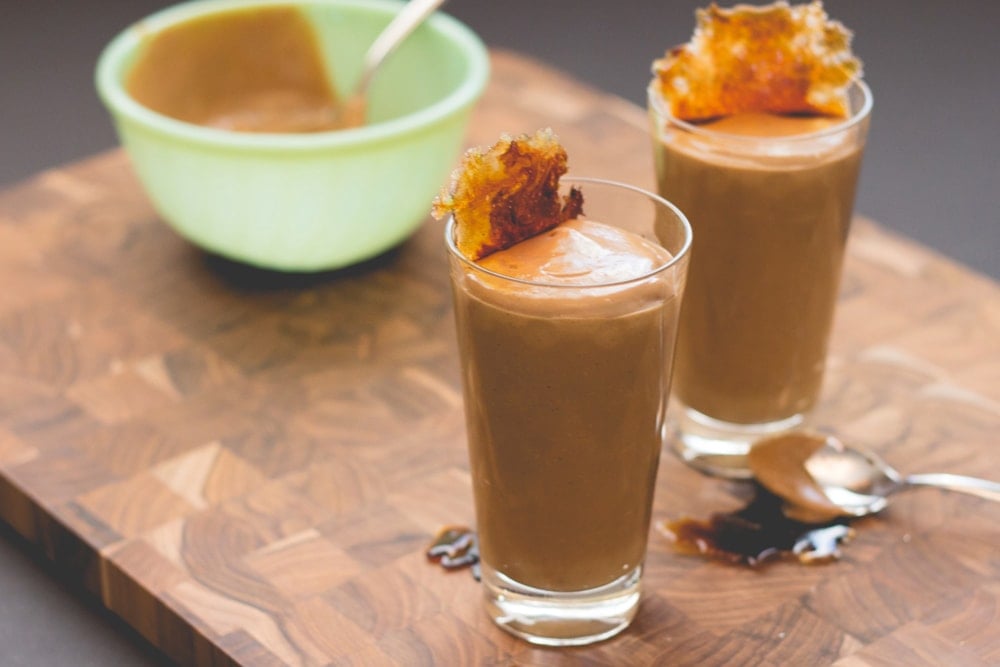 Tall glass filled with brown colored pudding with browned sugar crisp on top with light green bowl sitting in background all on wood cutting board surface