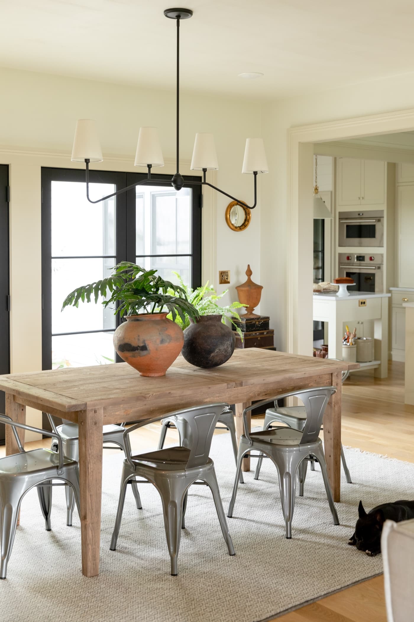 Wood dining table with steel chairs and white linen chandelier with black French doors in background