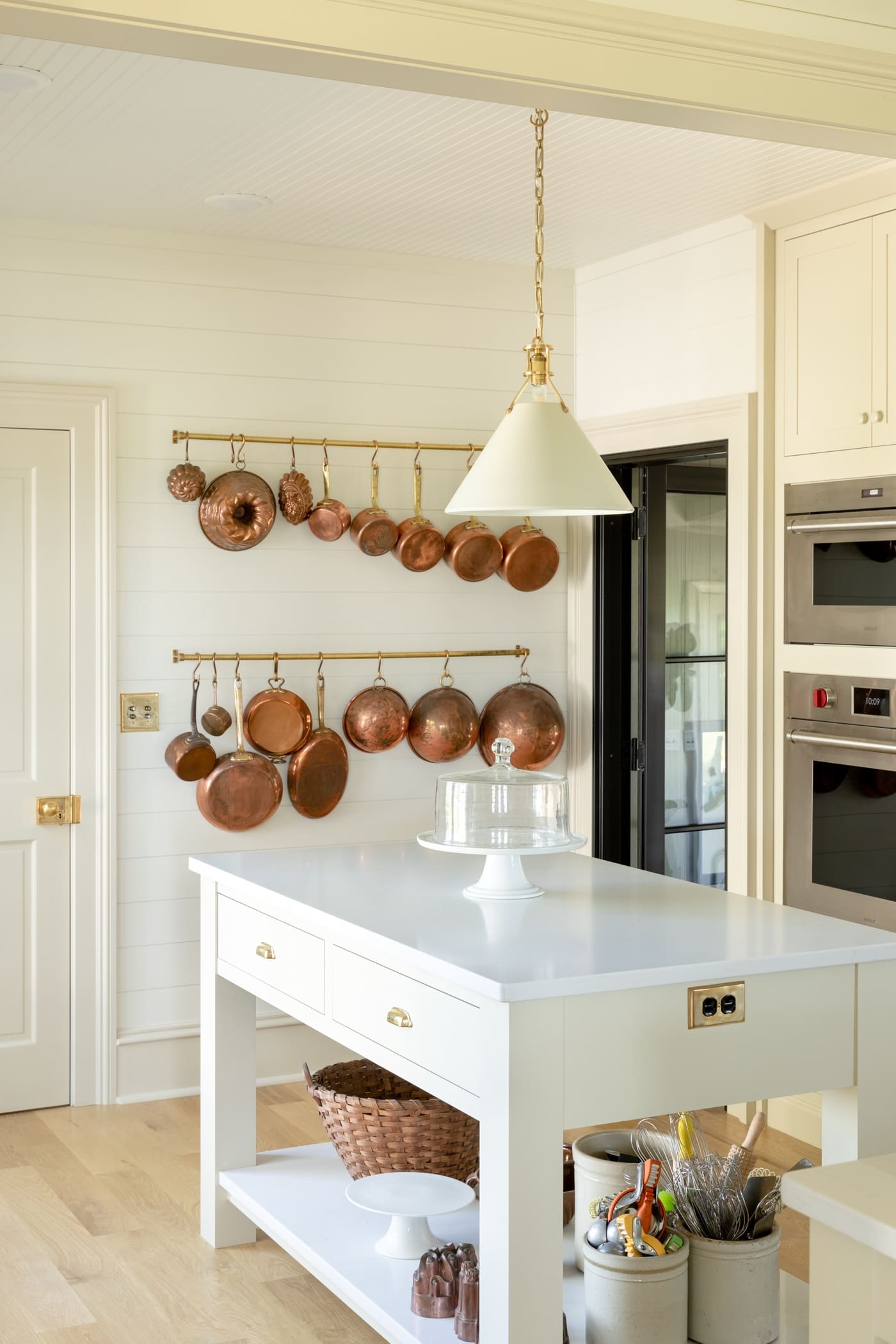 Small island with chunky legs and lower shelf covered in marble with wall oven behind and rack filled with copper cookware