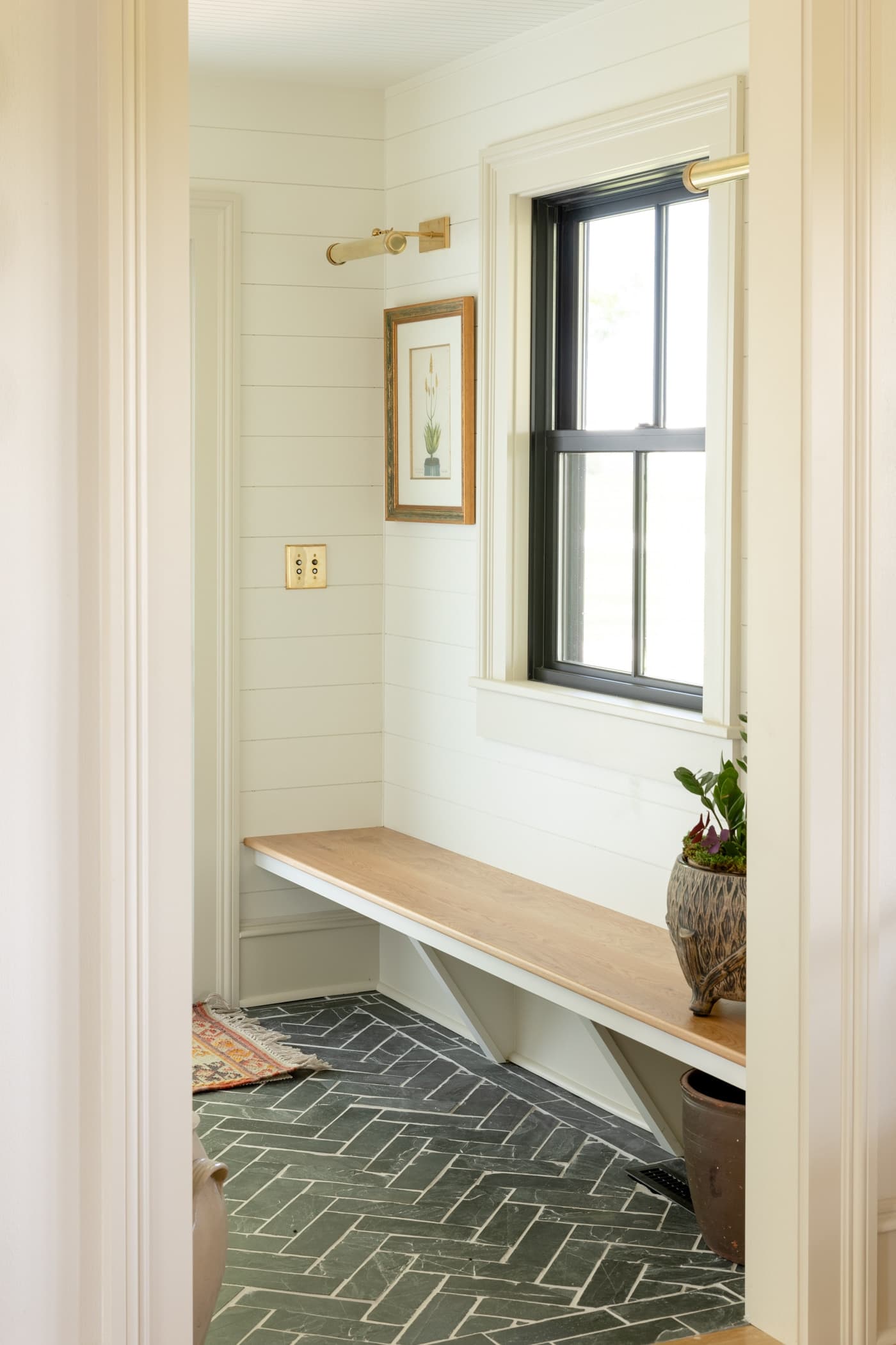 Dark gray slate floor and white oak bench make up a small entry space with black window and painting with a brass sconce above