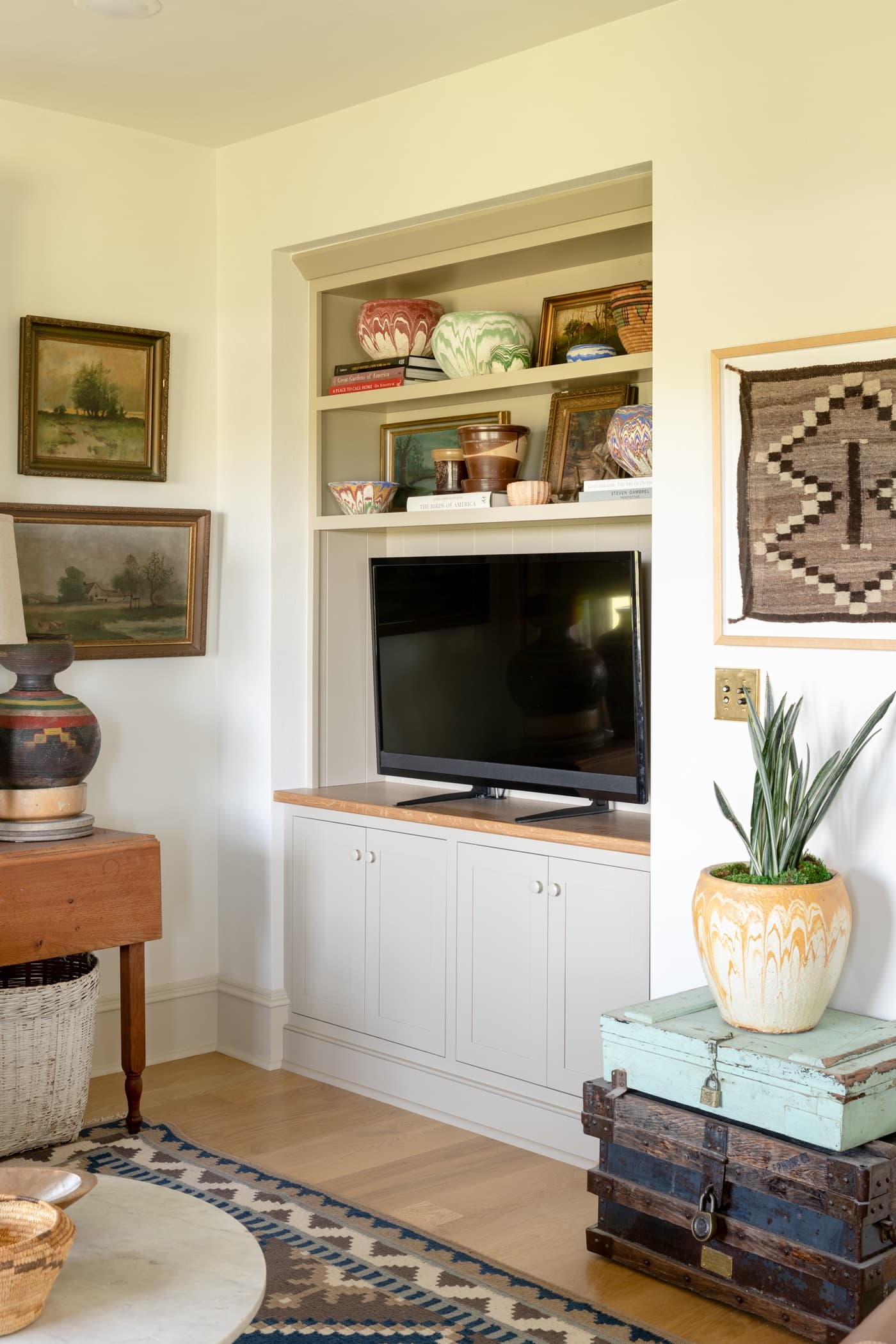 TV sitting in a built-in unit with cabinets underneath and shelves above with paintings on the surrounding walls and a blue rug on on the hardwood floors