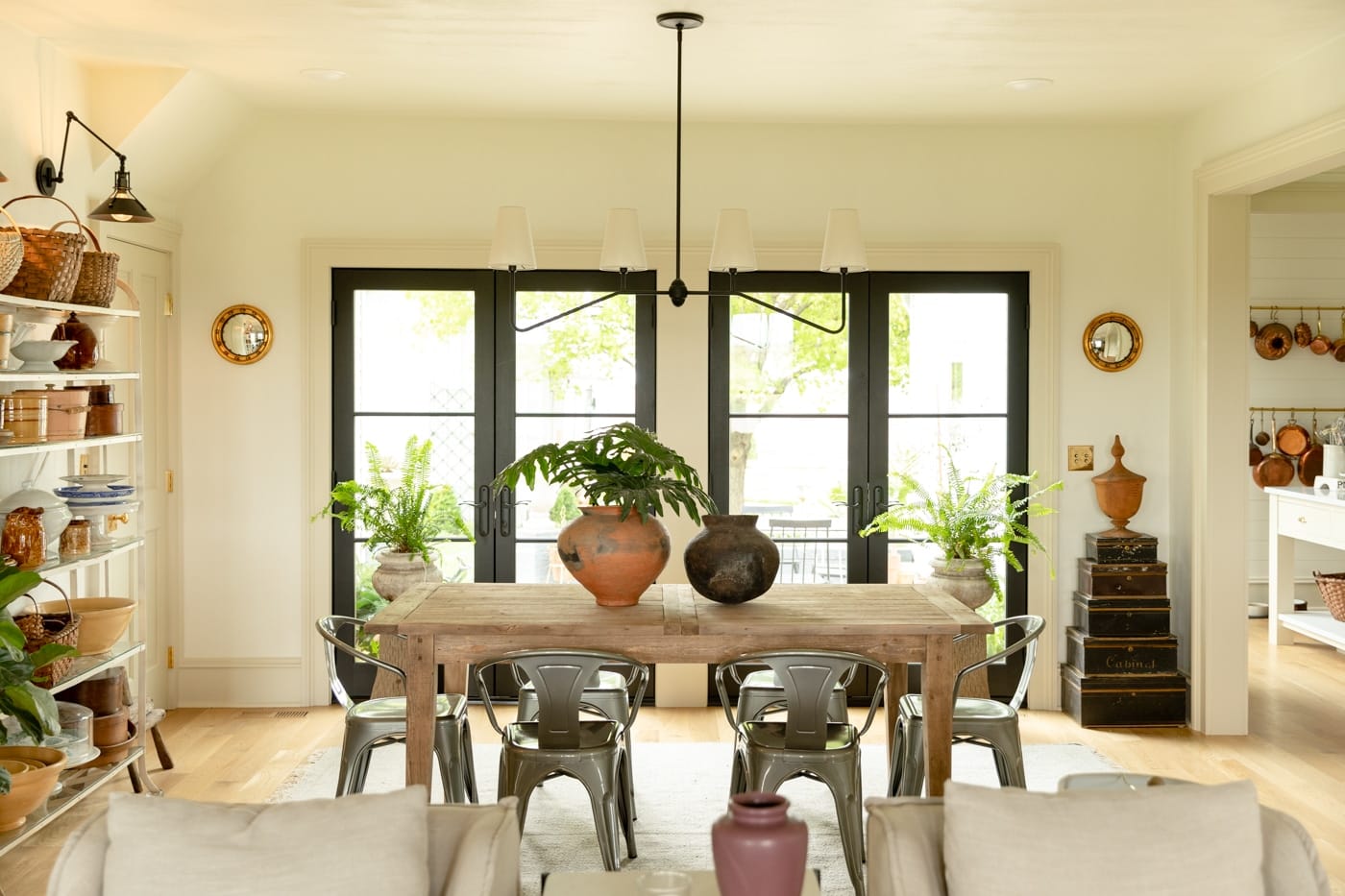 Wood dining room table with metal chairs around with pots and plants on table and large French doors in background