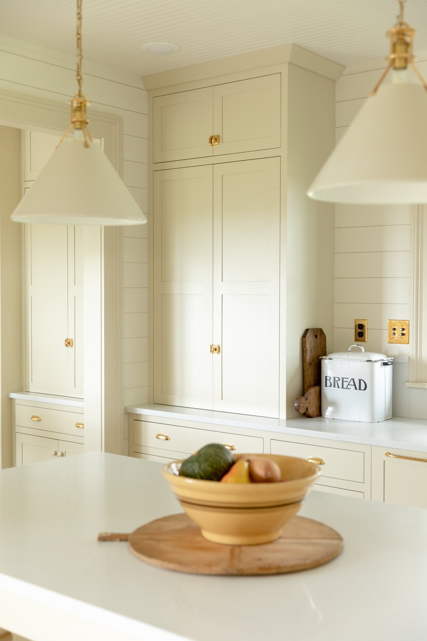Large pantry cabinet in creamy white farmhouse kitchen with yellow bowl filled with squash sitting on large island with white countertop and pendants hanging above island