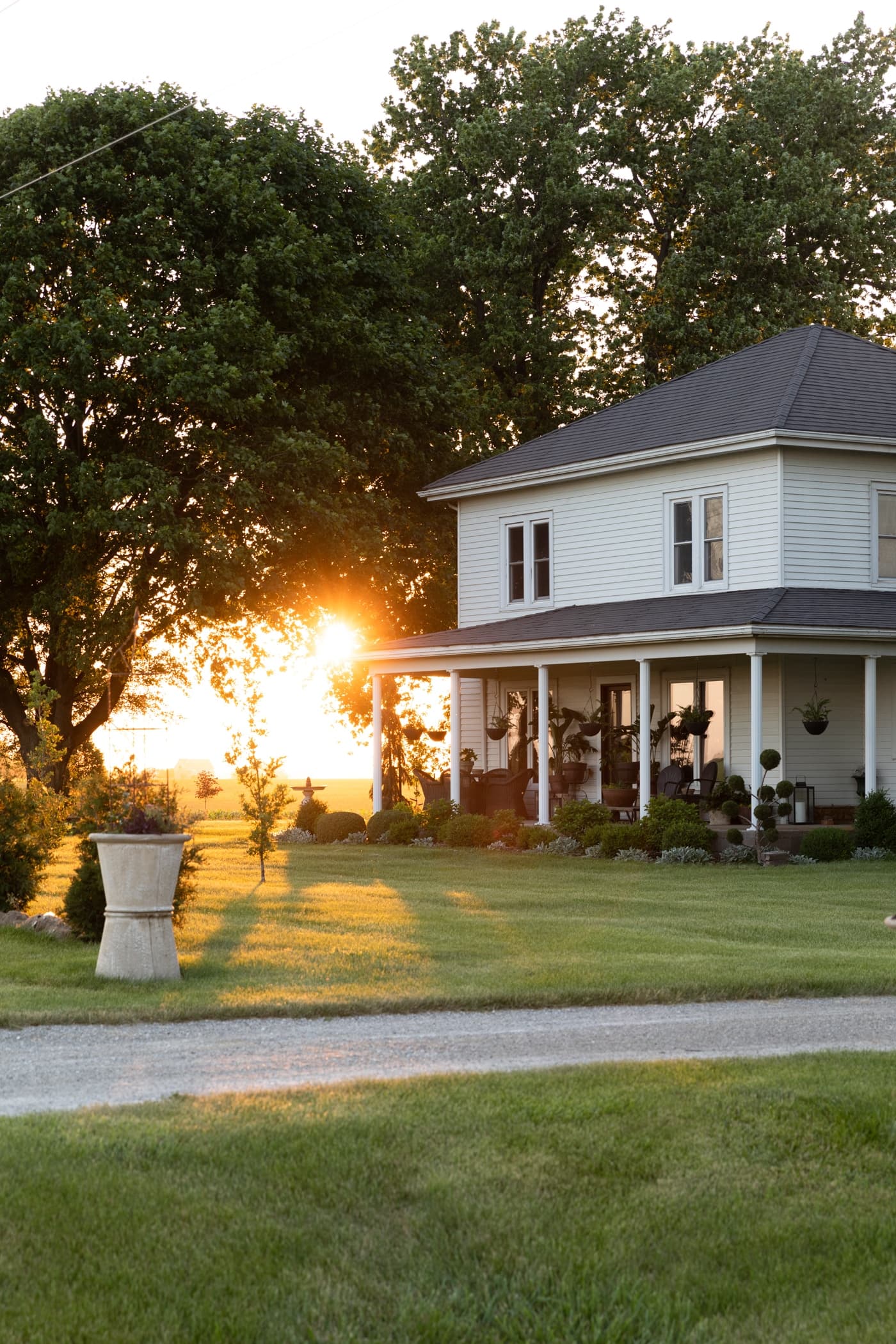 Exterior of white farmhouse with large front door and double windows with sun setting in background