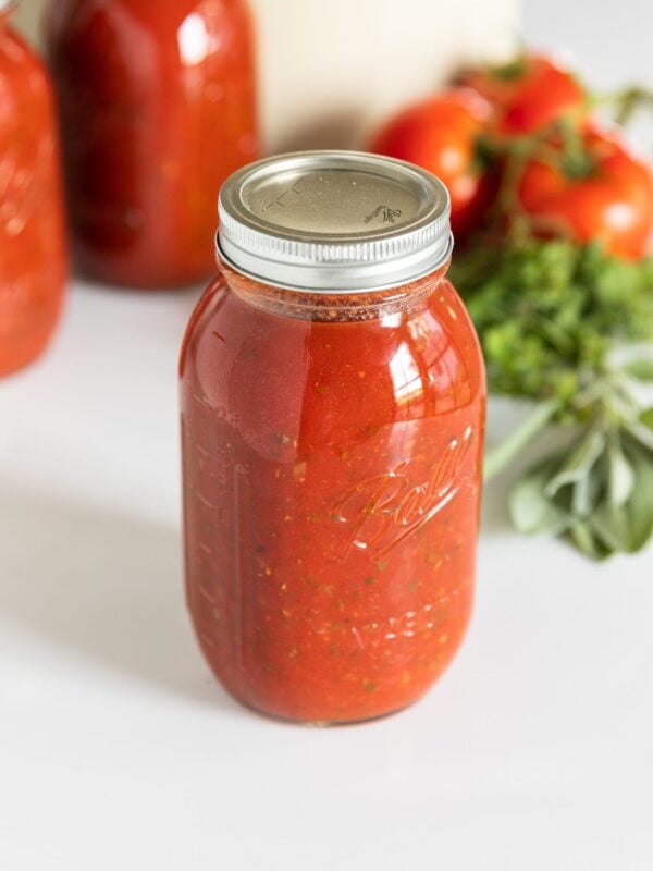Glass canning jar filled with red colored spaghetti sauce with lid and ring on top all on white countertop with herbs and tomatoes in background