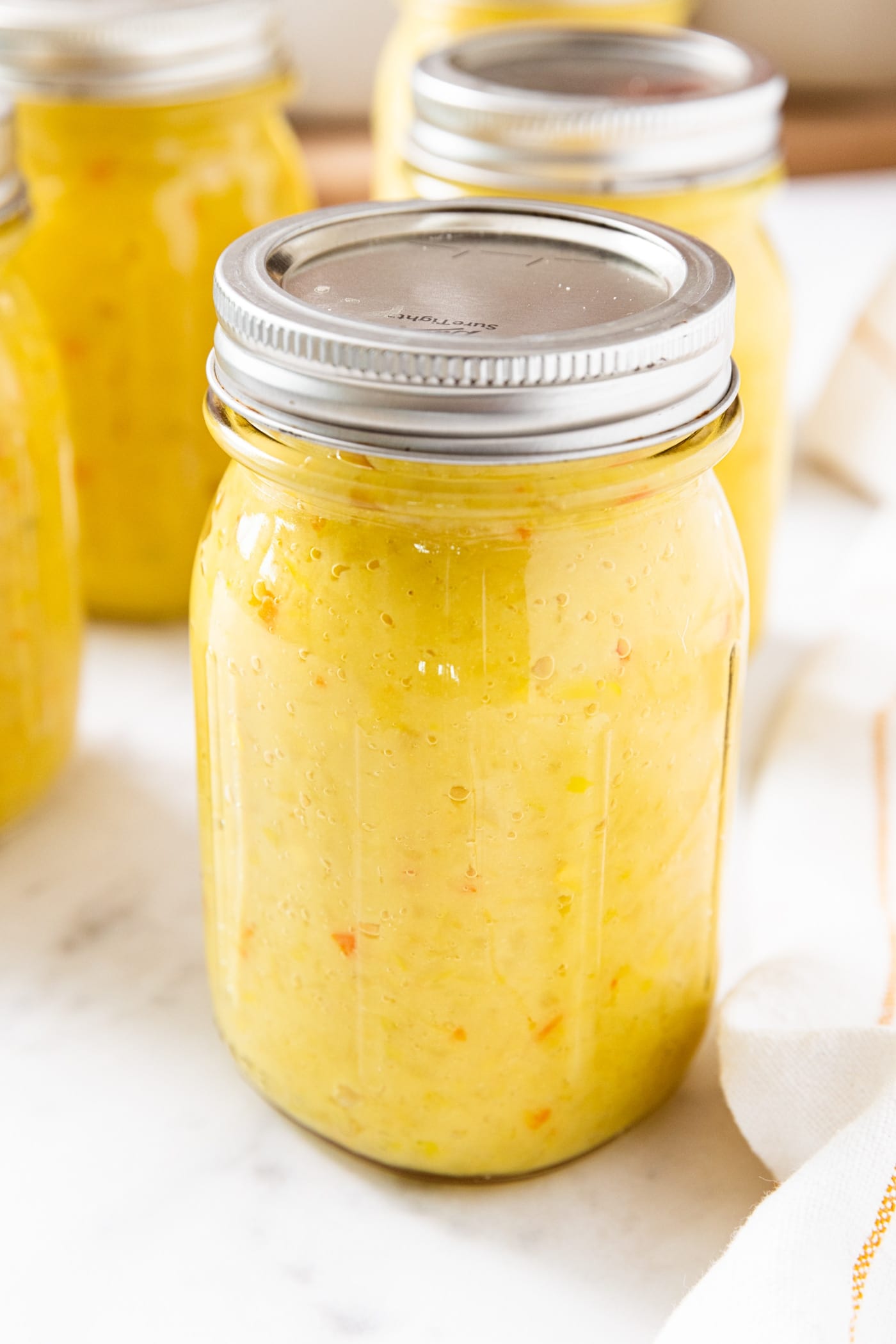 Glass canning jar filled with yellow colored hot pepper mustard sitting on marble surface with more jars in background.