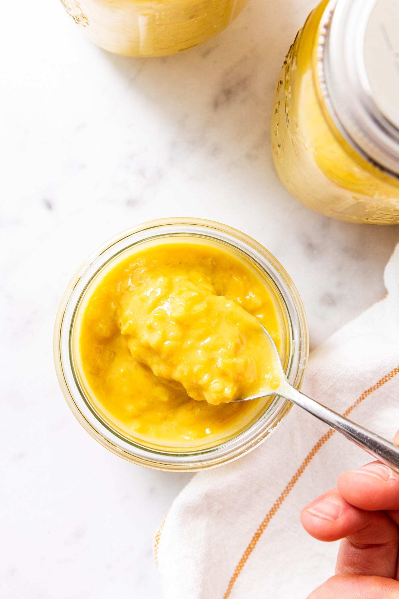 Hand holding metal spoon pulling some hot pepper mustard from small glass container sitting on marble surface with jars filled with canned mustard beside.