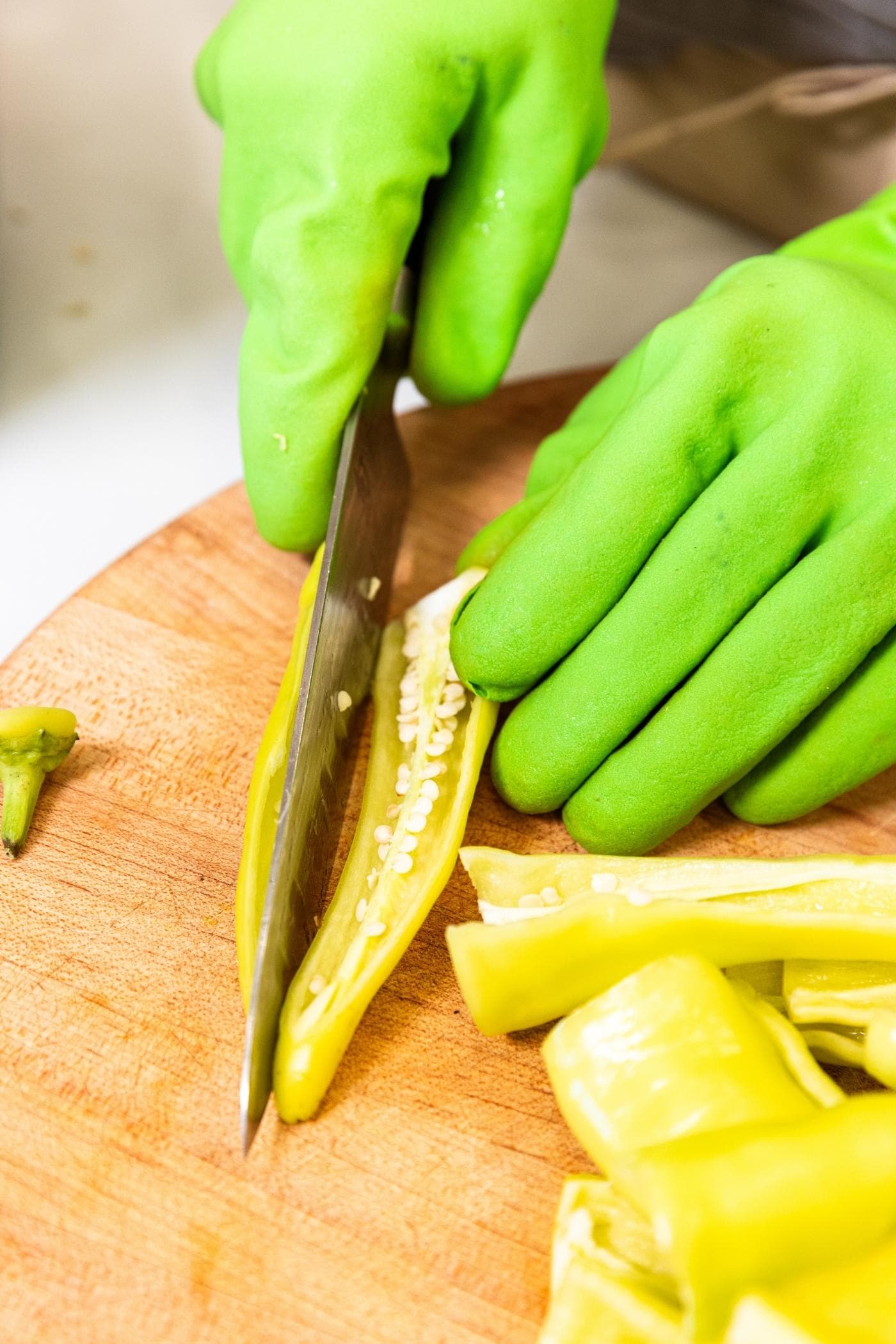 Hand in green plastic glove slicing yellow hot pepper in half on wood cutting board.