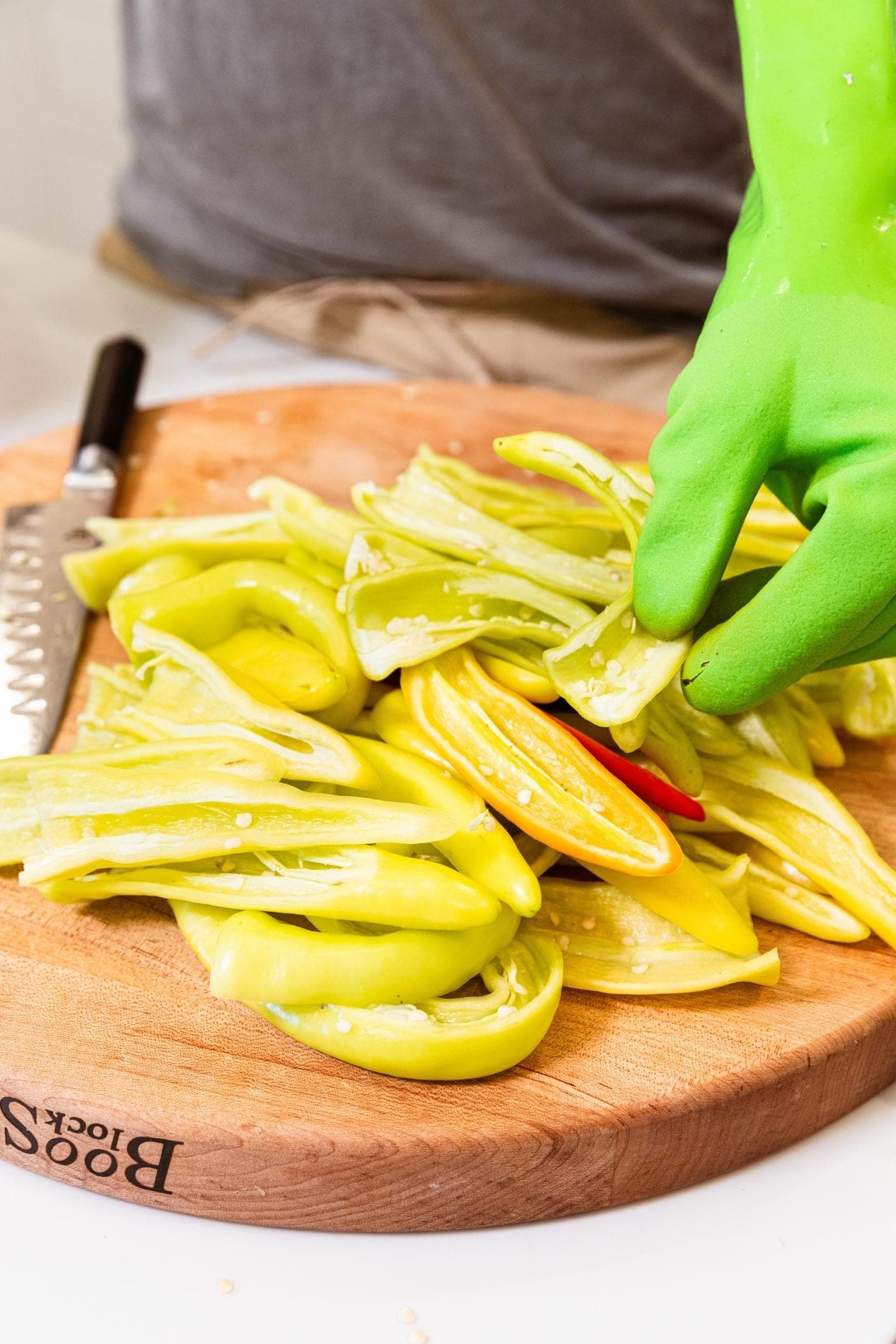 Hand in green plastic glove putting yellow hot pepper on a pile on wood cutting board.