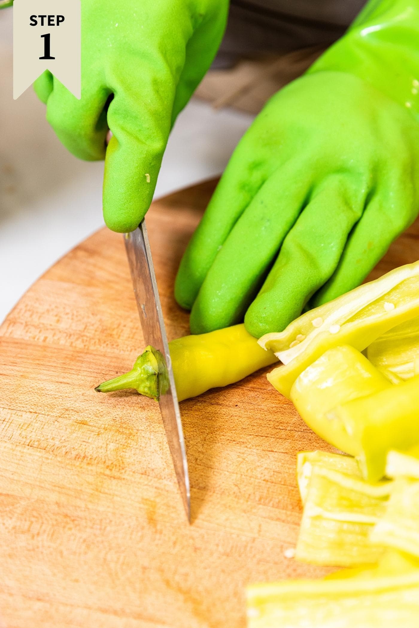 Hands in green plastic gloves cutting a yellow hot pepper on a wood cutting board.