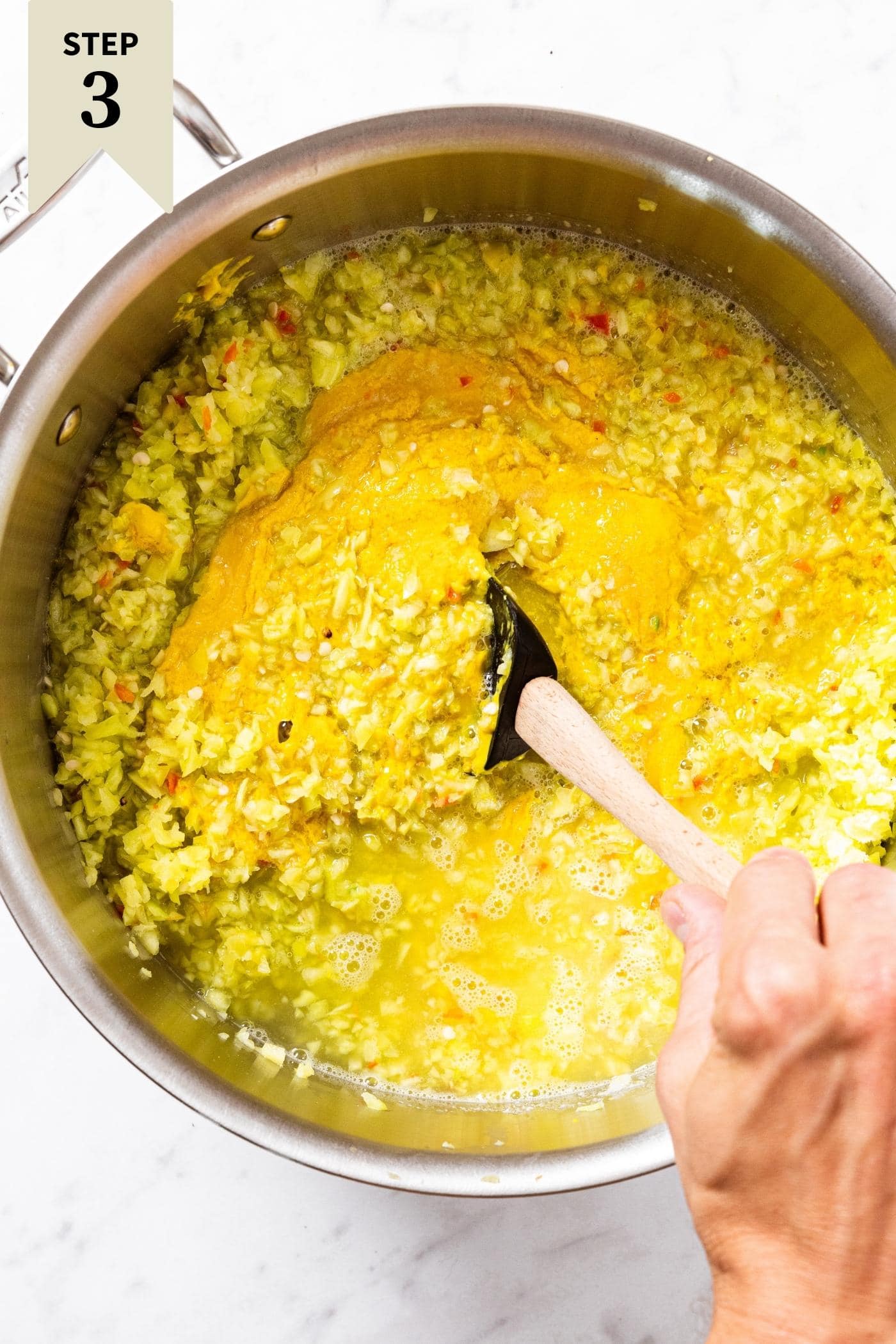 Hand stirring pepper and mustard mixture in large metal stockpot sitting on marble countertop.