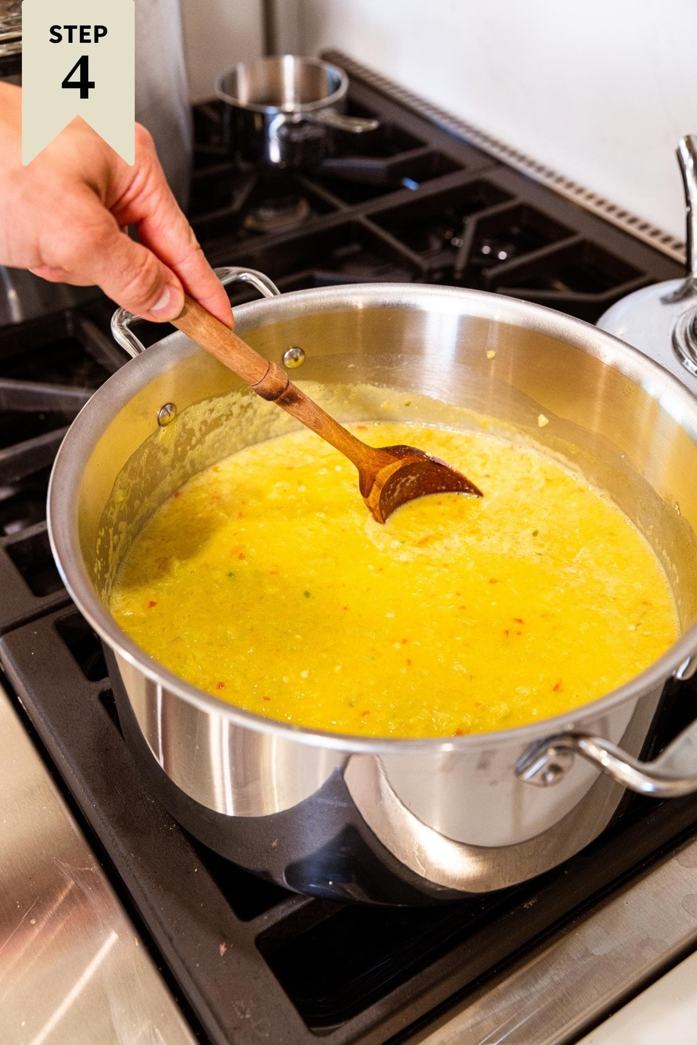 Hand holding wooden spoon stirring pepper mustard mixture in large metal stockpot on stove.