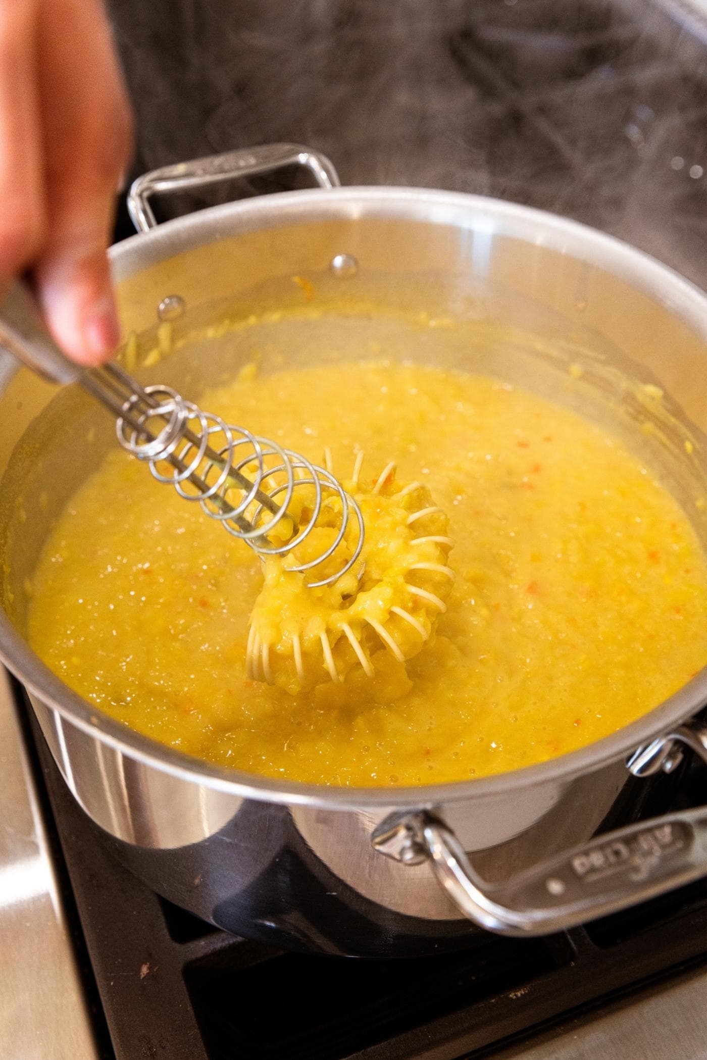 Hand holding whisk stirring mustard pepper mixture in large metal stockpot on stove.