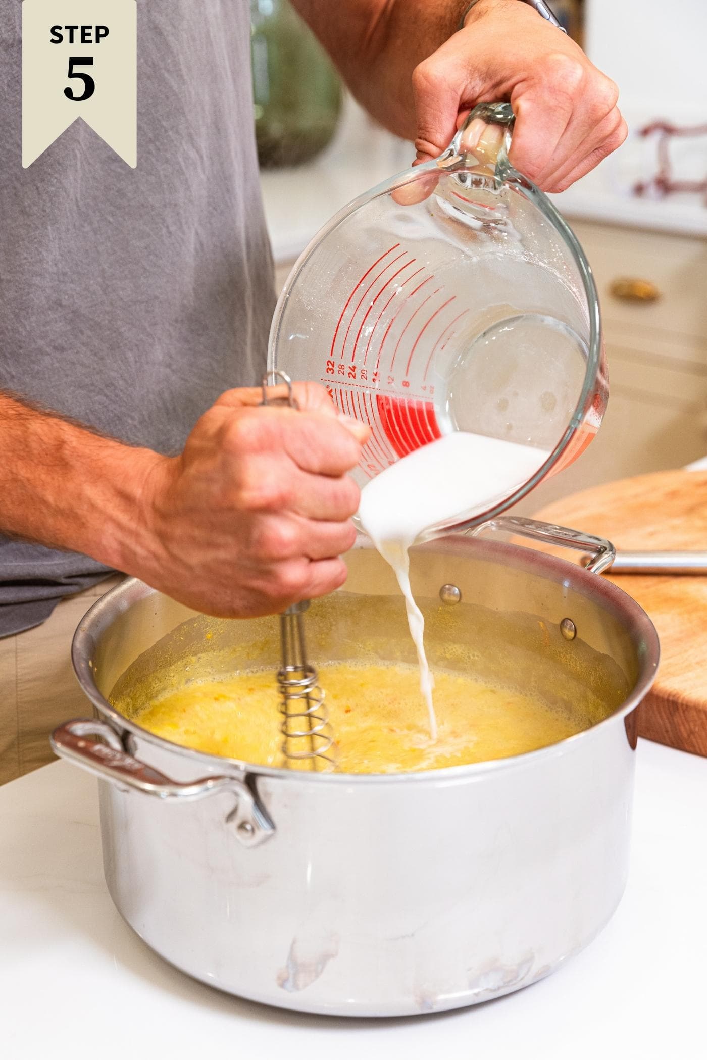 Hand holding measuring cup pouring white slurry mixture into large metal stockpot filled with yellow steaming mixture.